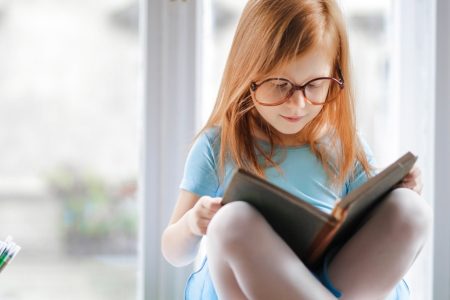 niña con gafas leyendo un libro cerca de la ventana de su casa