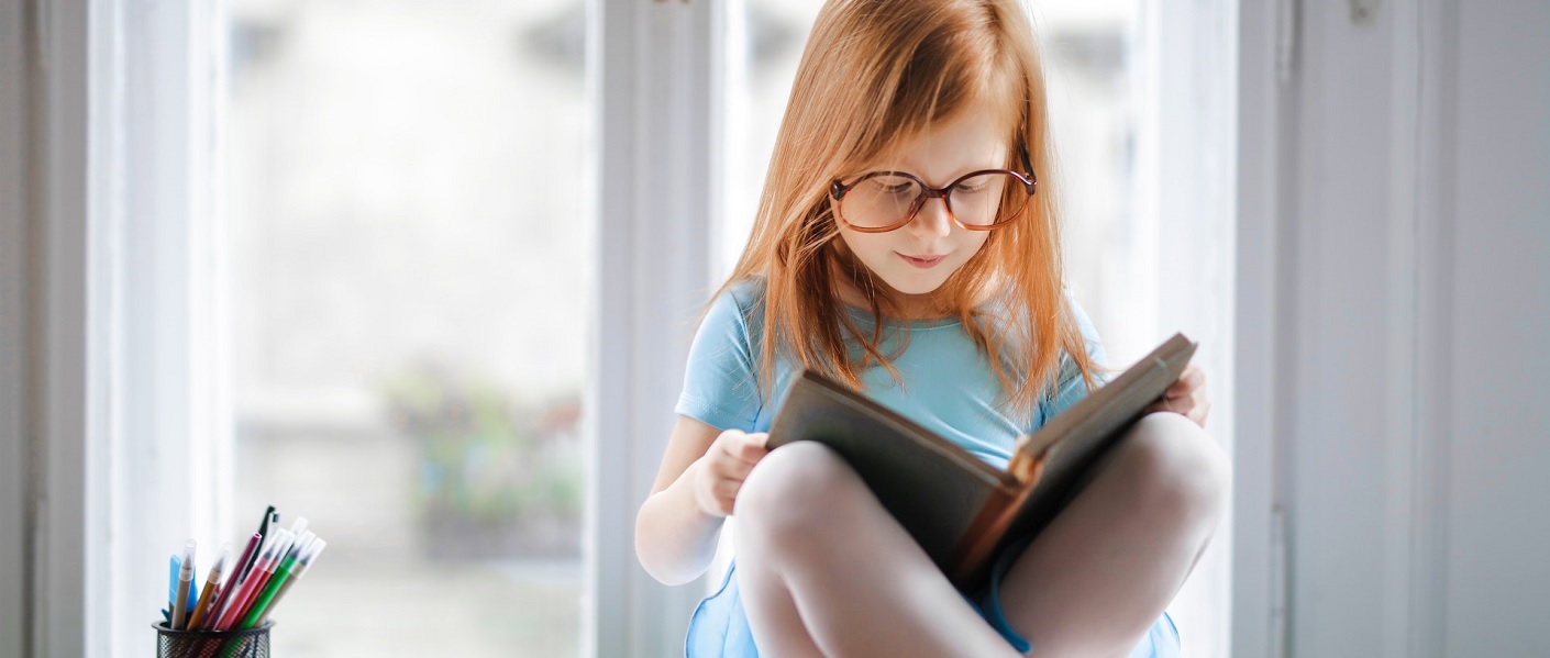 niña con gafas leyendo un libro cerca de la ventana de su casa