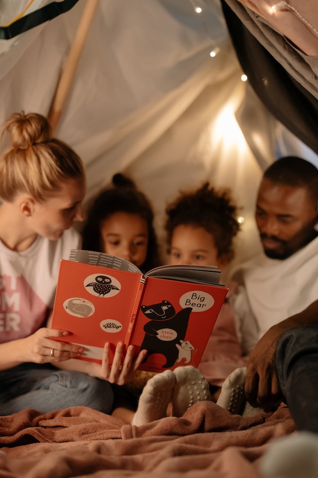 padres leyendo un cuento con sus hijos en una tienda de campaña improvisada en casa con sábanas antes de ir a dormir