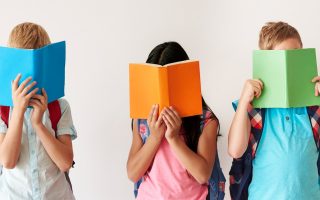 3 niños con mochila colgada en la espalda y tapándose la cara con un libro abierto cada uno