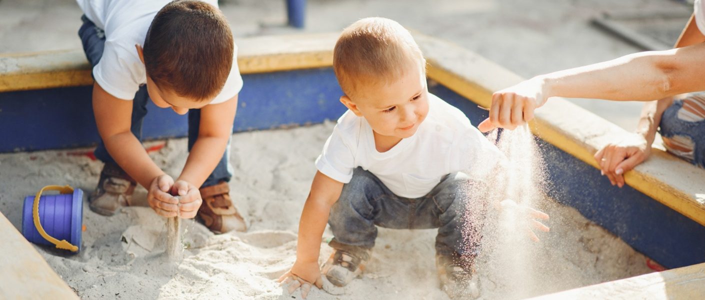 Dos niños pequeños jugando dentro de un arenero con su madre que agarra un puñado de arena con la mano que va soltando lentamente