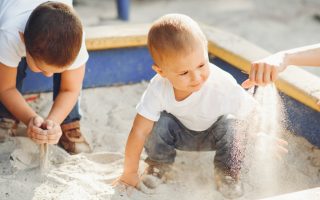 Dos niños pequeños jugando dentro de un arenero con su madre que agarra un puñado de arena con la mano que va soltando lentamente