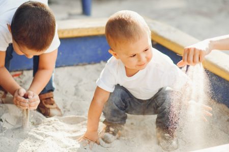 Dos niños pequeños jugando dentro de un arenero con su madre que agarra un puñado de arena con la mano que va soltando lentamente