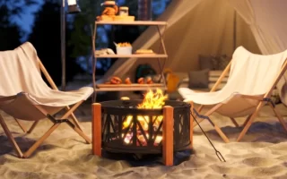 Brasero de leña con patas de madera en la playa junto a tienda de campaña y sillas plegables al atardecer