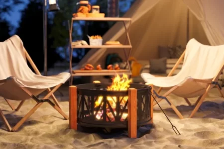 Brasero de leña con patas de madera en la playa junto a tienda de campaña y sillas plegables al atardecer