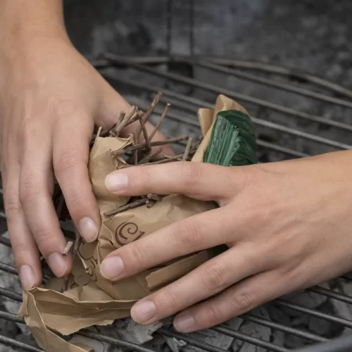 Manos colocando ramitas secas sobre papel para encender el carbón