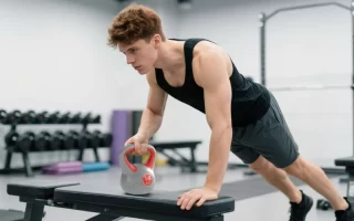 Hombre realizando plancha con remo usando una pesa rusa de 14 kg sobre un banco en el gimnasio 1400