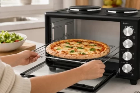 Persona sacando una rejilla con una pizza margarita recién hecha de un mini horno negro con dos placas de cocción encima. 1400